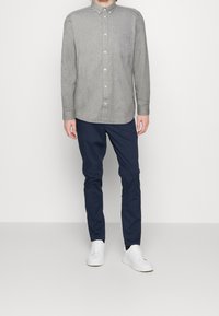 Chemise boutonnée à manches longues gris clair avec une poche sur la poitrine, associée à un pantalon slim-fit bleu marine et des baskets blanches. Design simple, texture lisse.
