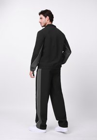 Μαύρο αθλητικό μπουφάν με γκρι μανίκια, ελαστικές μανσέτες και μαύρο παντελόνι που διαθέτει γκρι πλευρική ρίγα. Το μοντέλο στέκεται με την πλάτη γυρισμένη.