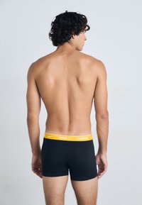 Boxers ajustados negros con una cinturilla amarilla de Nike. La tela parece suave y ceñida, con un diseño sencillo y sin patrones visibles.