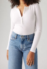 Eine Frau trägt ein weißes, geripptes Langarm-Henley-Shirt, das in hellblaue, hochgeschnittene Jeans gesteckt ist, und steht vor einem schlichten Hintergrund.