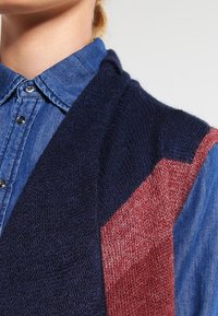 Närbild av en person som bär en blå denimskjorta med knappar under en marinblå och rostfärgad stickad väst.