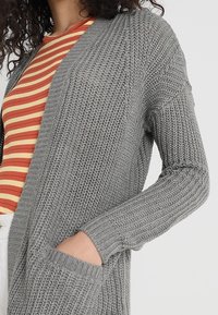 Mujer vistiendo un cárdigan de punto gris con un bolsillo frontal sobre una camiseta a rayas roja y amarilla, con la mano colocada de manera casual en el bolsillo.