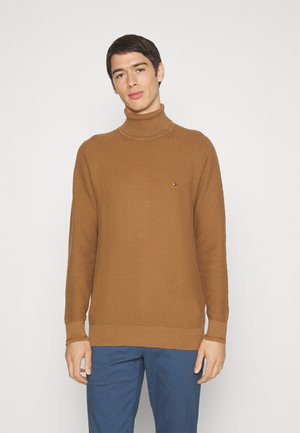 Maglione a collo alto marrone con texture, bordi a coste nelle maniche e nell'orlo, con un piccolo logo sul petto. Indossato con pantaloni blu.
