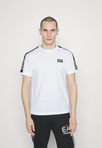T-shirt en coton blanc à manches courtes, avec des accents noirs et un logo sur la poitrine. Le design inclut des rayures à motifs sur les épaules.