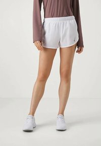 Pantalones cortos deportivos blancos de Nike con una cintura elástica, combinados con una camiseta de manga larga marrón, contra un fondo neutro.