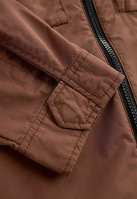 Manche de veste marron avec détail de poignet cousu et fermeture éclair noire sur fond de tissu lisse.