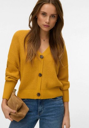 Sennepsgul strikket cardigan med V-udskæring, tre brune knapper og opkørte ærmer. Stylet med blå jeans og en beige clutch.