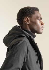 Chaqueta negra con capucha, de textura suave, cierre con cremallera y cordones ajustables. Cuello alto con un aspecto superpuesto sobre un fondo neutro.