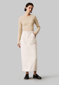Beige hosszú ujjú felső, fehér farmer maxi szoknyával párosítva, amelynek két elülső zsebe és gombos záródása van. A modellen fekete cipő van.