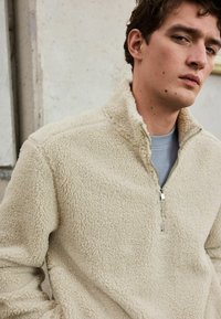 Pull-over en polaire beige avec un col zippé mi-hauteur, extérieur texturé et intérieur doux. Présente un design minimaliste et des détails de couture subtils.