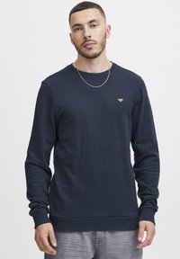 Mornarsko modra trenirka iz mehkega materiala s krožnim izrezom, rebrastimi manšeti in robom, ki ima na prsih majhen trikotni logotip.