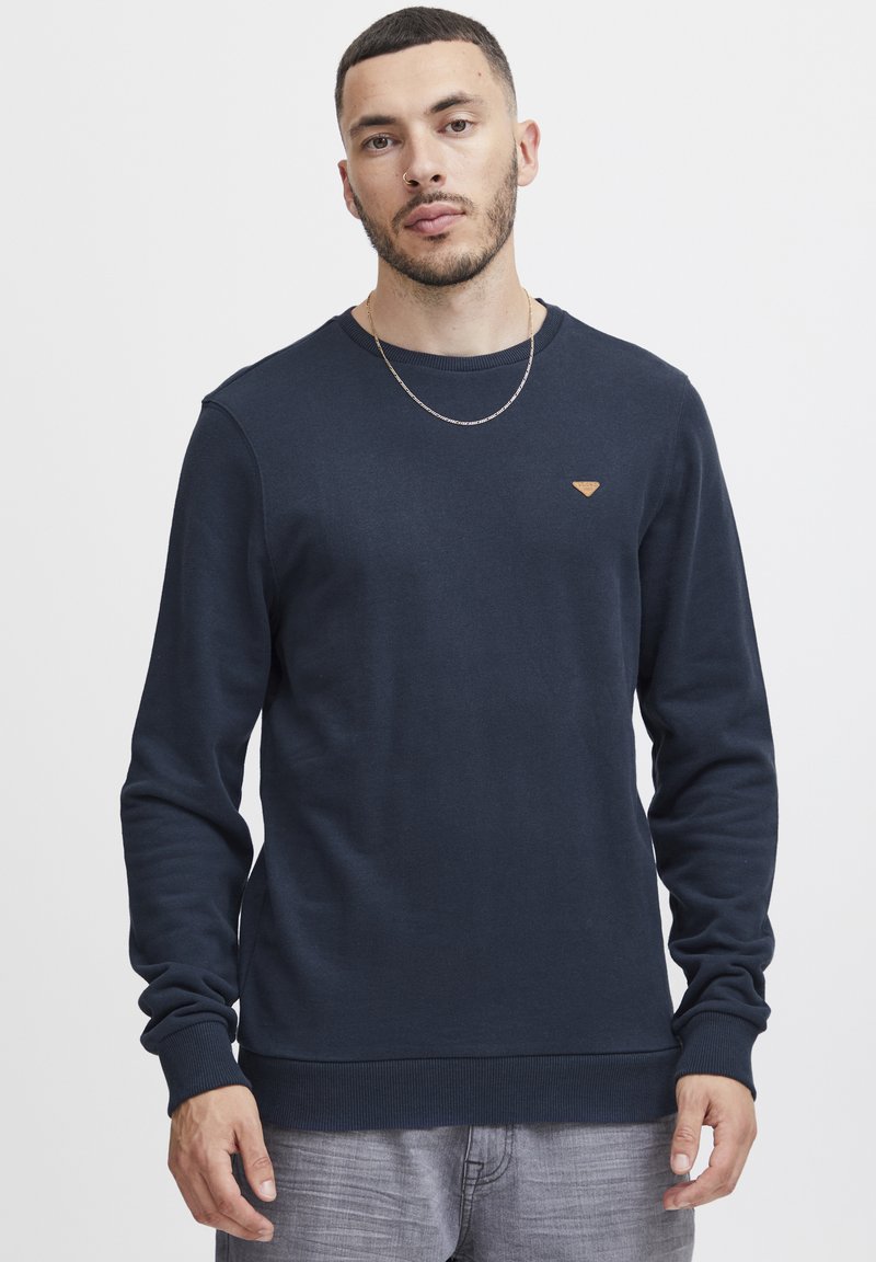 Mornarsko modra trenirka iz mehkega materiala s krožnim izrezom, rebrastimi manšeti in robom, ki ima na prsih majhen trikotni logotip.