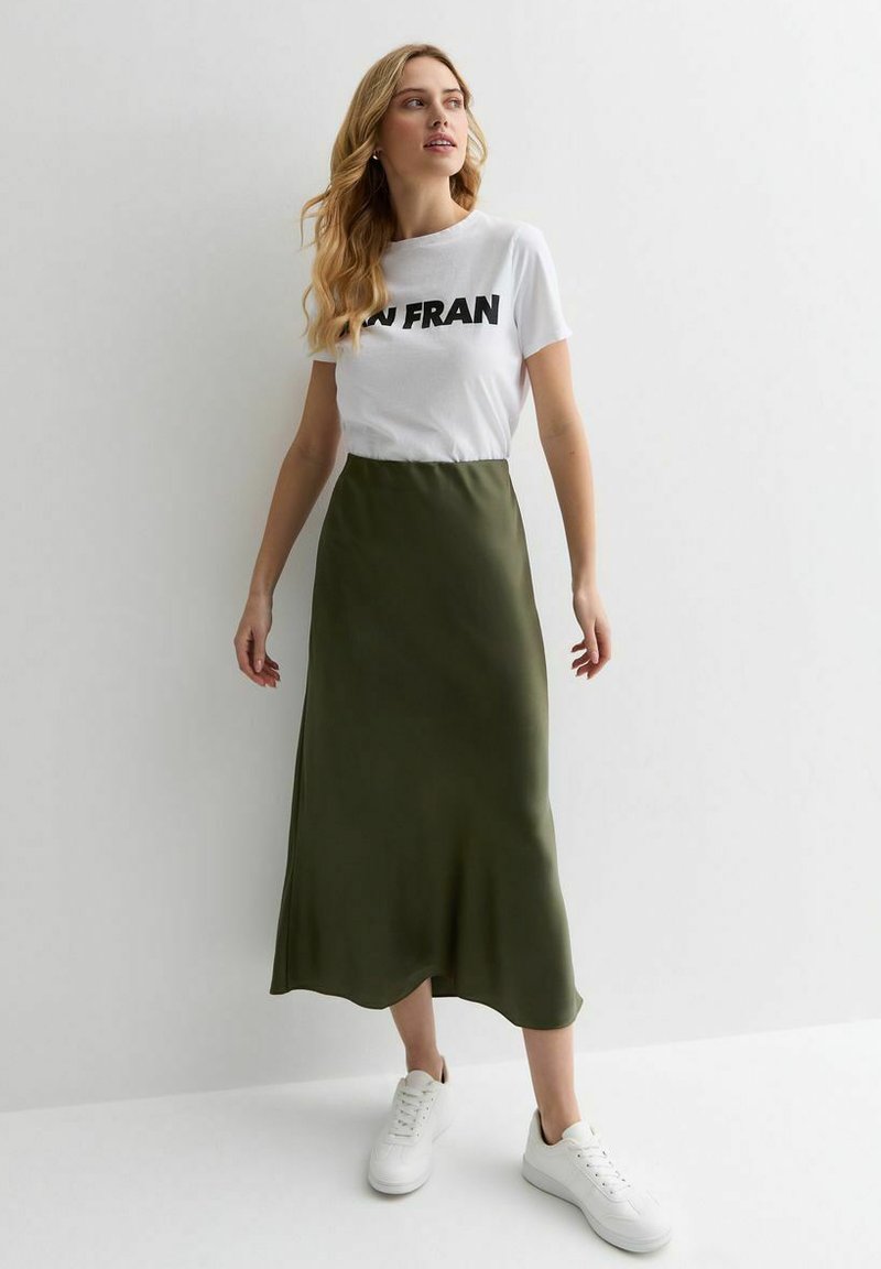 Witte T-shirt met zwarte tekst gecombineerd met een lange olijfgroene satijnen rok, aangevuld met witte sneakers. Eenvoudige, relaxte pasvorm en vloeiend ontwerp.