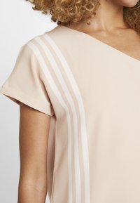 Svetle béžová krátká rukávová blůza s výstřihem do V, která má dvě bílé svislé pruhy podél ramena a jemnou, hladkou texturu.