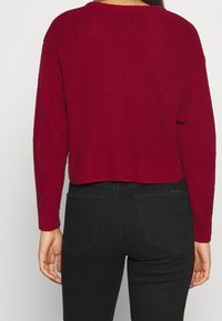 Viininpunainen neulepaita, jossa on cropped-muotoilu, ribbauskudos ja pitkät hihat, yhdistettynä mustiin muotoutuviin farkkuihin, joissa on takataskut.