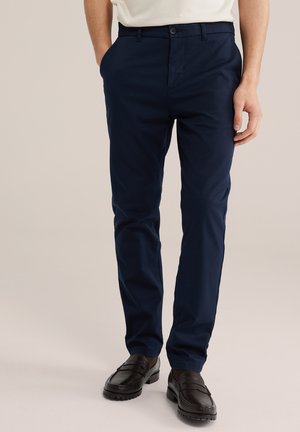 Man draagt marineblauwe slim-fit chino's met zijzakken en zwarte leren loafers, staand tegen een neutrale achtergrond.
