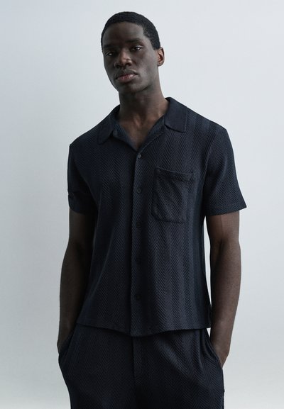 Homme portant une chemise à manches courtes sombre boutonnée et un pantalon assorti, debout les mains dans les poches devant un fond clair uni.