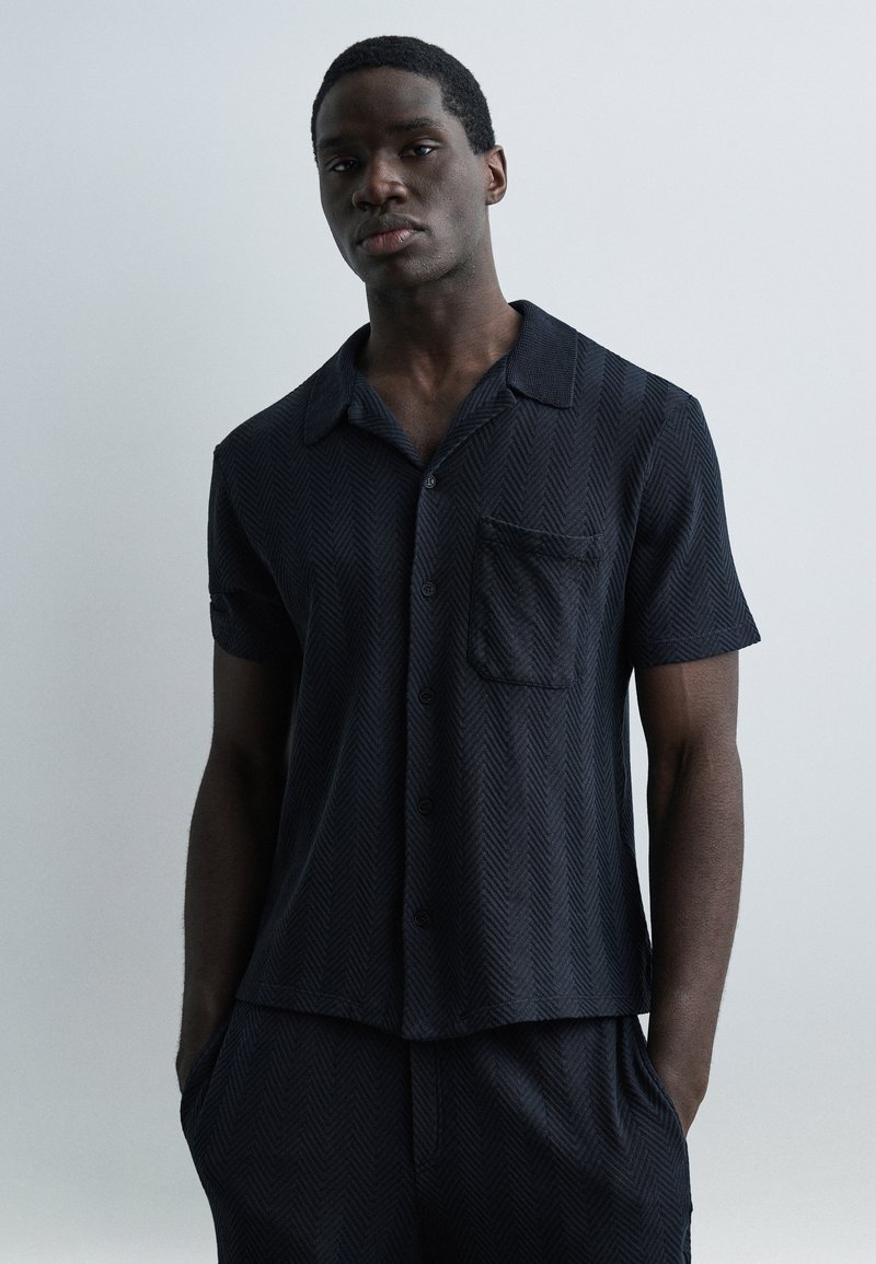 Homme portant une chemise à manches courtes sombre boutonnée et un pantalon assorti, debout les mains dans les poches devant un fond clair uni.