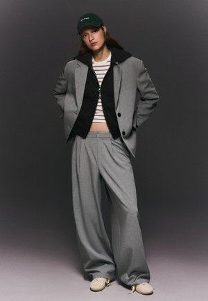 Giovane donna che indossa un ampio completo grigio, felpa nera con zip, top corto a righe, cappellino verde con scritta e sneakers beige, in piedi con le mani dietro.