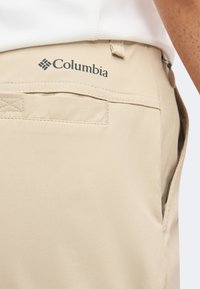 Μπεζ παντελόνι Columbia με πίσω τσέπη και λογότυπο στη μέση, φορεμένο με λευκό πουκάμισο μέσα από το παντελόνι.