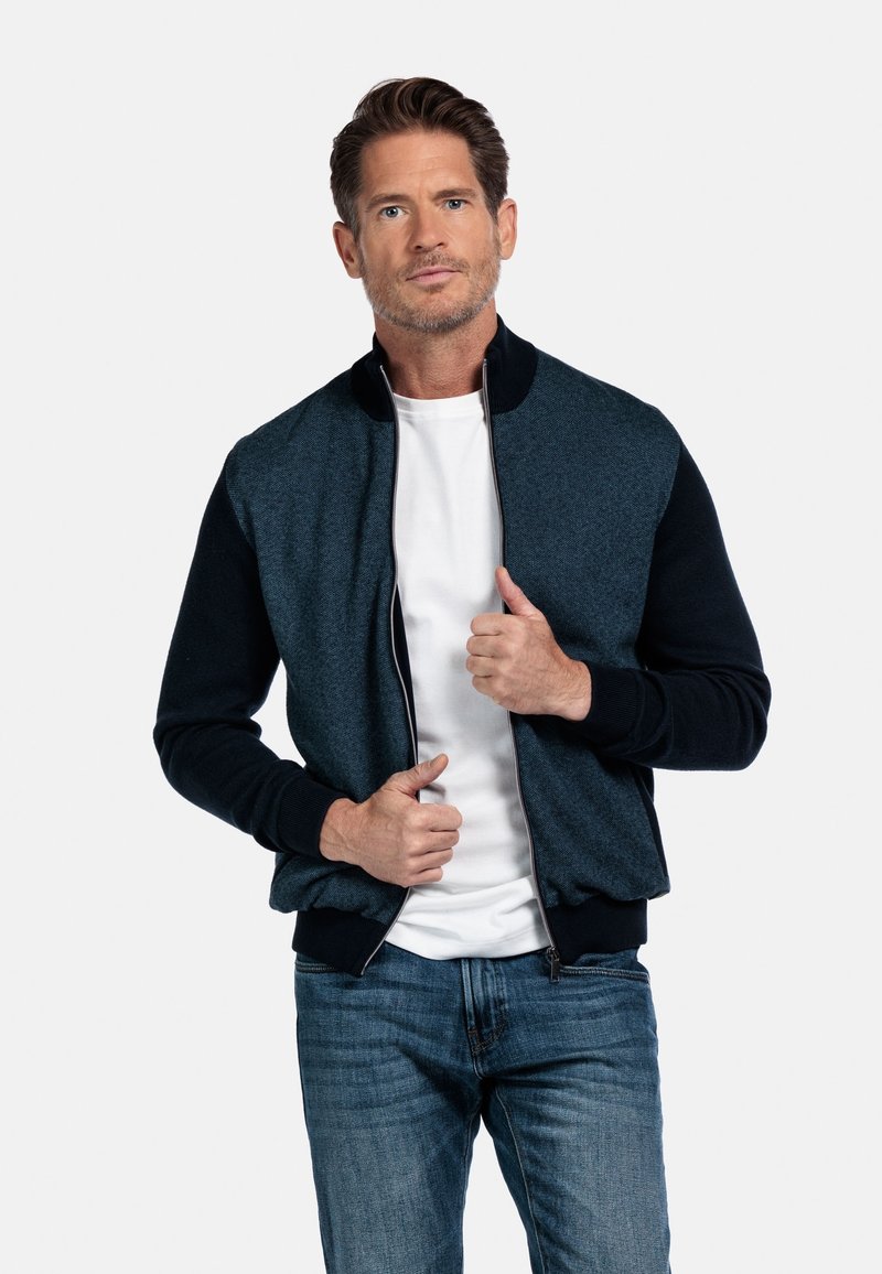 Bomberjack in donkerblauw en zwart, met een rits aan de voorkant, geribde kraag en manchetten, en textuurstof. Gedragen over een witte t-shirt.