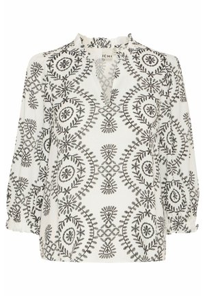 Blouse blanche avec broderies florales noires, dotée d'un col volanté, de manches longues et d'une coupe décontractée. Motifs circulaires notables dans l'ensemble.