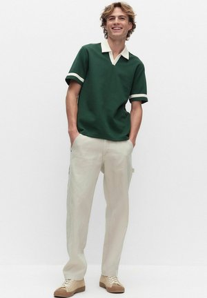 Hymyilevä nuori mies kiharat hiukset, vihreä poolopaita valkoisella kauluksella ja kalvosimilla, beigeit housut ja beiget tennarit, kädet taskuissa.