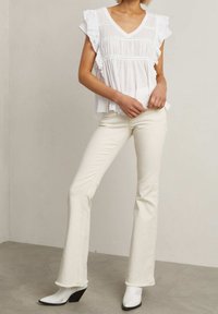 Vit rynkad blus med V-ringning och texturerade detaljer, i kombination med vita utsvängda jeans och spetsiga vita klackstövlar.