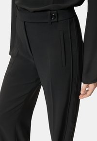 Pantaloni neri su misura in tessuto strutturato, caratterizzati da una texture liscia, tasche laterali, chiusura con bottone e dettagli plissettati.