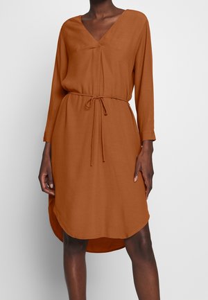 Femme portant une robe à manches longues, couleur rouille, longueur au genou, avec un col en V et une ceinture nouée à la taille.