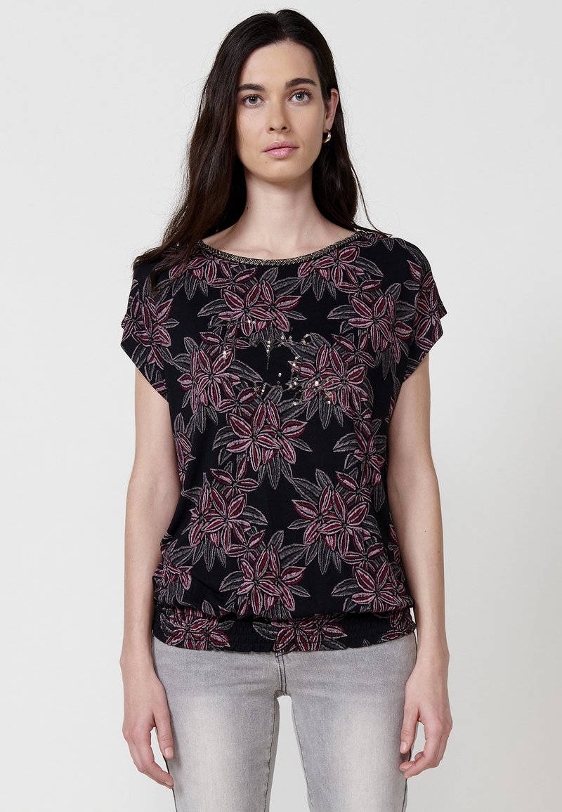 Chemise noire à motifs floraux en rose et gris, à manches courtes et avec un bas froncé. Tissu lisse avec un léger brillant.
