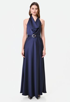 Frau trägt ein bodenlanges marineblaues Satin-Halterkleid mit drapiertem Ausschnitt und einem Taillengürtel mit dekorativer Schnalle.