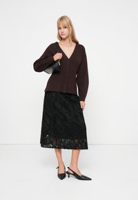 Σκούρο μπορντό ζακέτα με κουμπιά και V-λαιμόκοψη, συνδυασμένη με μια μαύρη μίντι φούστα με υφή κυματιστών σχεδίων. Μαύρες γόβες slingback ολοκληρώνουν την εμφάνιση.