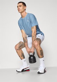 Blauw gestreept kort mouwen shirt, witte shorts, witte sport schoenen met rode accenten. Persoon heft een zwarte kettlebell op en toont tattoos.