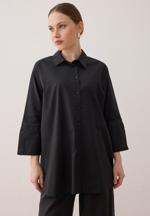 Donna dalla pelle chiara e capelli raccolti che indossa una camicia nera ampia con bottoni e polsini larghi, posando davanti a uno sfondo chiaro uniforme.