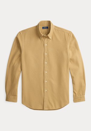 Camisa de manga larga color mostaza hecha de tela suave. Presenta un cuello con botones, botones frontales y un pequeño logo bordado en el pecho.