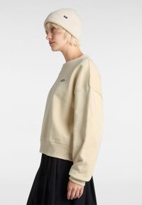 Sudadera beige con dobladillo y puños acanalados, que cuenta con un logo bordado en el pecho. Complementa una falda negra con pliegues y un gorro de lana.