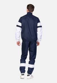 Tuta da ginnastica blu navy con accenti bianchi. La giacca presenta polsini e vita elasticizzati; i pantaloni hanno righe abbinate e orli elasticizzati.