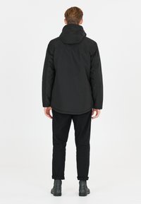 Zwarte waterdichte jack met een capuchon, lange mouwen en een rechte snit, gecombineerd met zwarte broeken en enkellaarsjes. Gladde textuur.