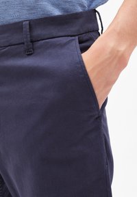 Ruka odložená v kapse tmavě modrých kalhot, ukazující strukturovanou modrou košili zasunutou dovnitř, na bílém pozadí.