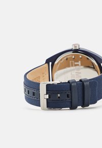 Correa de reloj de textil azul marino con detalles en cuero, hebilla de metal plateado y caja redonda de acero inoxidable. Parte trasera etiquetada con el nombre de la marca.
