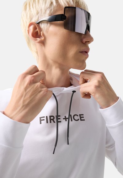 Person with short blonde hair wearing large black sunglasses and a white hoodie with the text "FIRE+ICE," pulling at the hood strings.