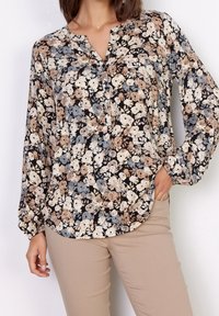 Bloes met bloemenprint op een zwarte achtergrond, met beige, blauwe en witte bloemen. Lange mouwen en een knoopsluiting bij de hals voegen detail toe.