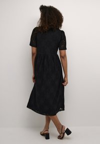 Zwarte kanten jurk met korte pofmouwen, een aansluitend lijfje en een vloeiende rok met geschulpte zoomdetails. Draagt met sandalen op blokhak.