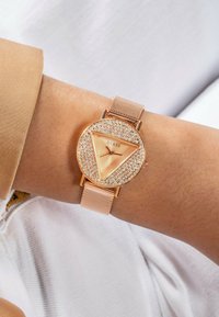 Orologio oro rosa con una faccia triangolare, circondata da cristalli scintillanti. Il bracciale a rete completa il design elegante. Caratterizzato dal marchio GUESS.
