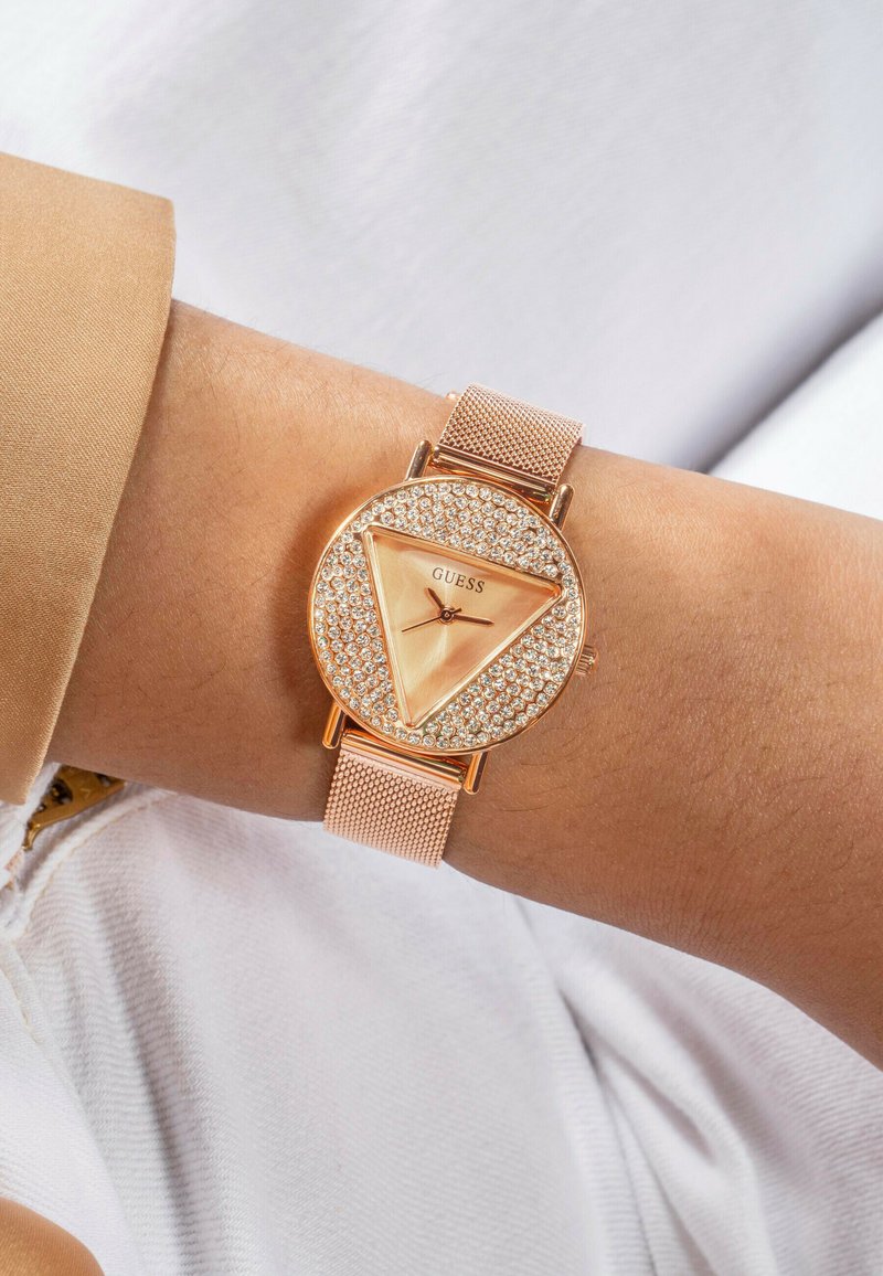 Orologio oro rosa con una faccia triangolare, circondata da cristalli scintillanti. Il bracciale a rete completa il design elegante. Caratterizzato dal marchio GUESS.
