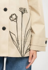 Giacca beige con ricamo floreale nero sulla tasca, con due bottoni neri e una vestibilità comoda. Jeans blu visibili sotto.