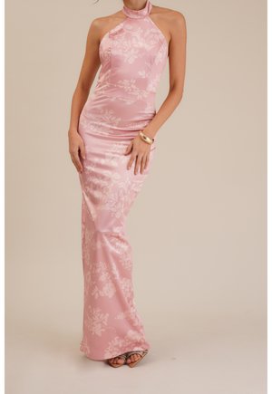 Kobieta w dopasowanej różowej sukni z atłasu na szyję z kwiatowymi wzorami, złotą bransoletką i butami na obcasie z odkrytymi palcami, stojąca na jednolitym tle.