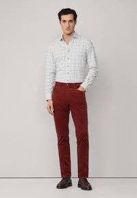 Pantaloni in velluto a coste rossi, camicia a quadri bianca e blu con bottoni in legno. L'outfit è abbinato a scarpe stringate marroni scure.