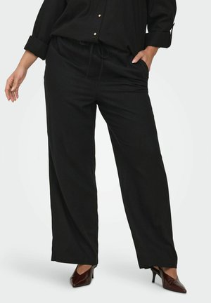 Vrouw die losse zwarte broek met trekkoord, zwart blouse met opgerolde mouwen en bruine pumps met puntige neus draagt, staand met hand in zak.